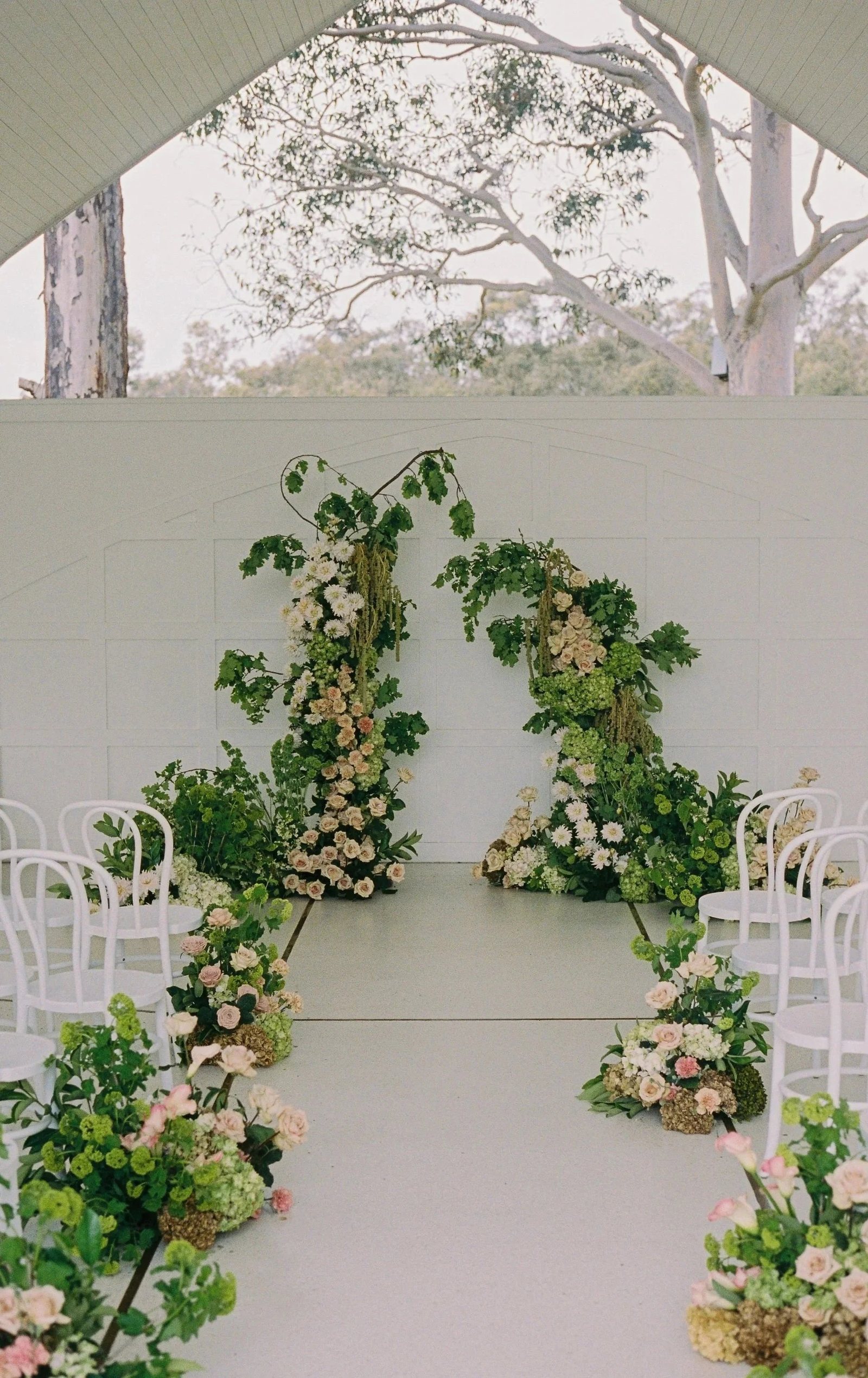 The ceremony aisle set with white chairs and two floral pillars under the gums
